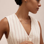 A woman with dark hair in a neat bun wears a sleeveless, cream top with red stripes and the Colette Droop Link Huggie Earrings in Silver, along with a simple necklace, bracelet, and ring, looking to the side against a light background.