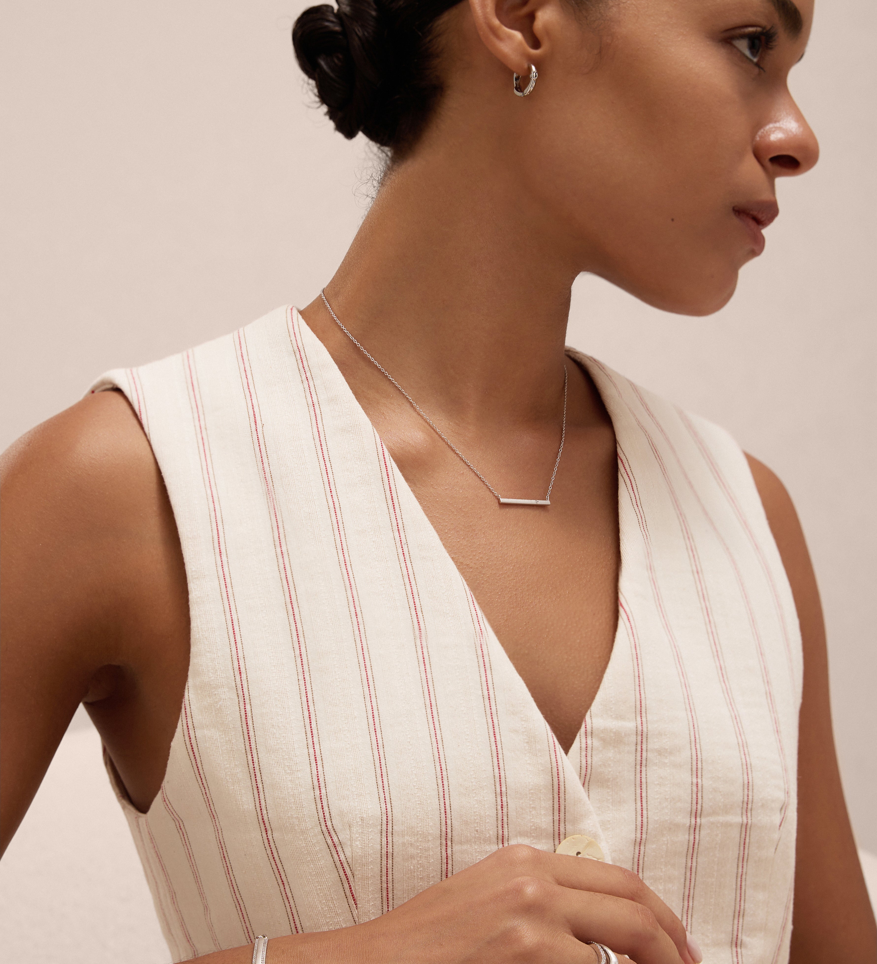 A woman with dark hair in a neat bun wears a sleeveless, cream top with red stripes and the Colette Droop Link Huggie Earrings in Silver, along with a simple necklace, bracelet, and ring, looking to the side against a light background.