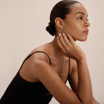 A woman with slicked-back hair wears a black sleeveless top and the Colette Droop Link Huggie Earrings in Silver, along with silver rings, a necklace, and a bracelet. She gazes thoughtfully to the side with her chin resting on her hand.