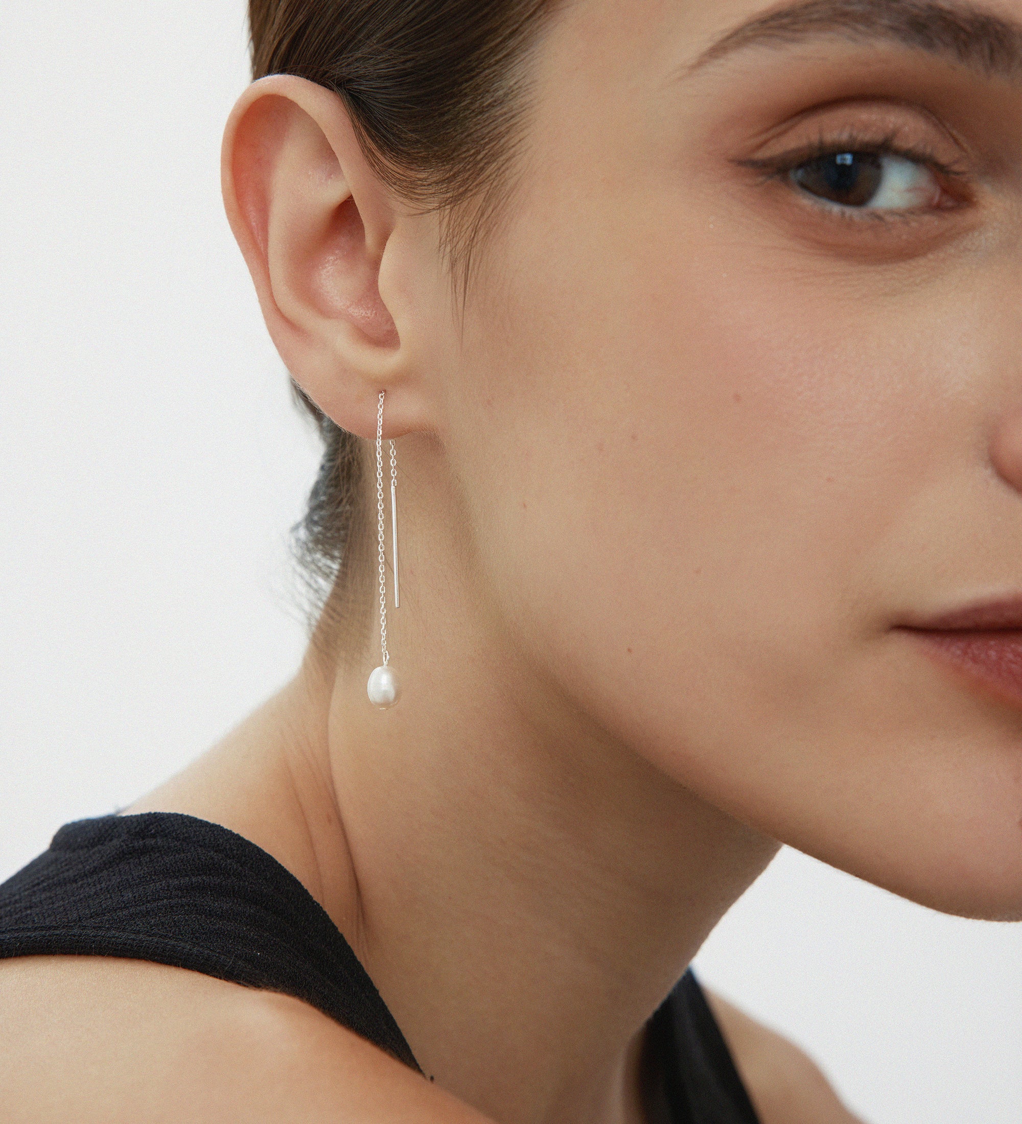 Close-up of a woman with brown hair in a black top, wearing Eira Pearl Drop Threader Earrings in Silver. She faces slightly right, highlighting her cheek, lips, and the elegant shimmer of her pearl drop earrings.