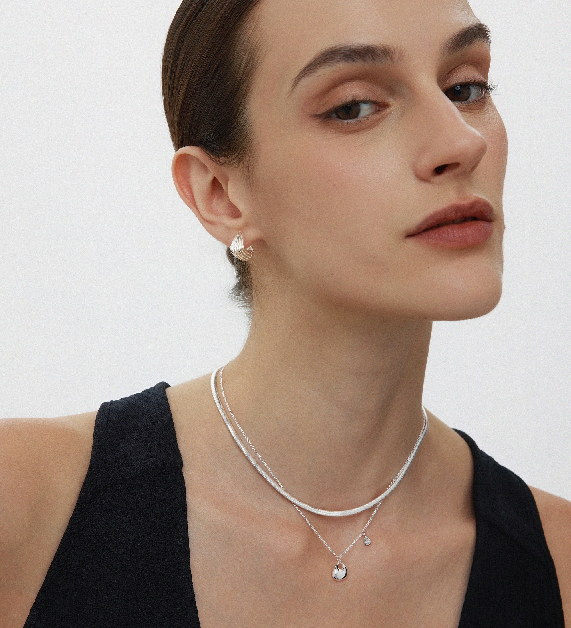 A woman with brown hair in a sleek updo wears the Estelle Herringbone Chain Necklace in Silver layered with another pendant and matching silver studs, paired with a black sleeveless top against a plain white background.