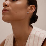 A woman with smooth skin and dark hair in a low bun wears a sleeveless cream top with red stripes, showcasing the Regina Horizontal Bar Pendant Necklace in Silver and minimalist triangle earrings as she gazes upward calmly.