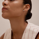 A woman with smooth skin and dark hair in a low bun wears a sleeveless cream top with red stripes, showcasing the Regina Horizontal Bar Pendant Necklace in Silver and minimalist triangle earrings as she gazes upward calmly.