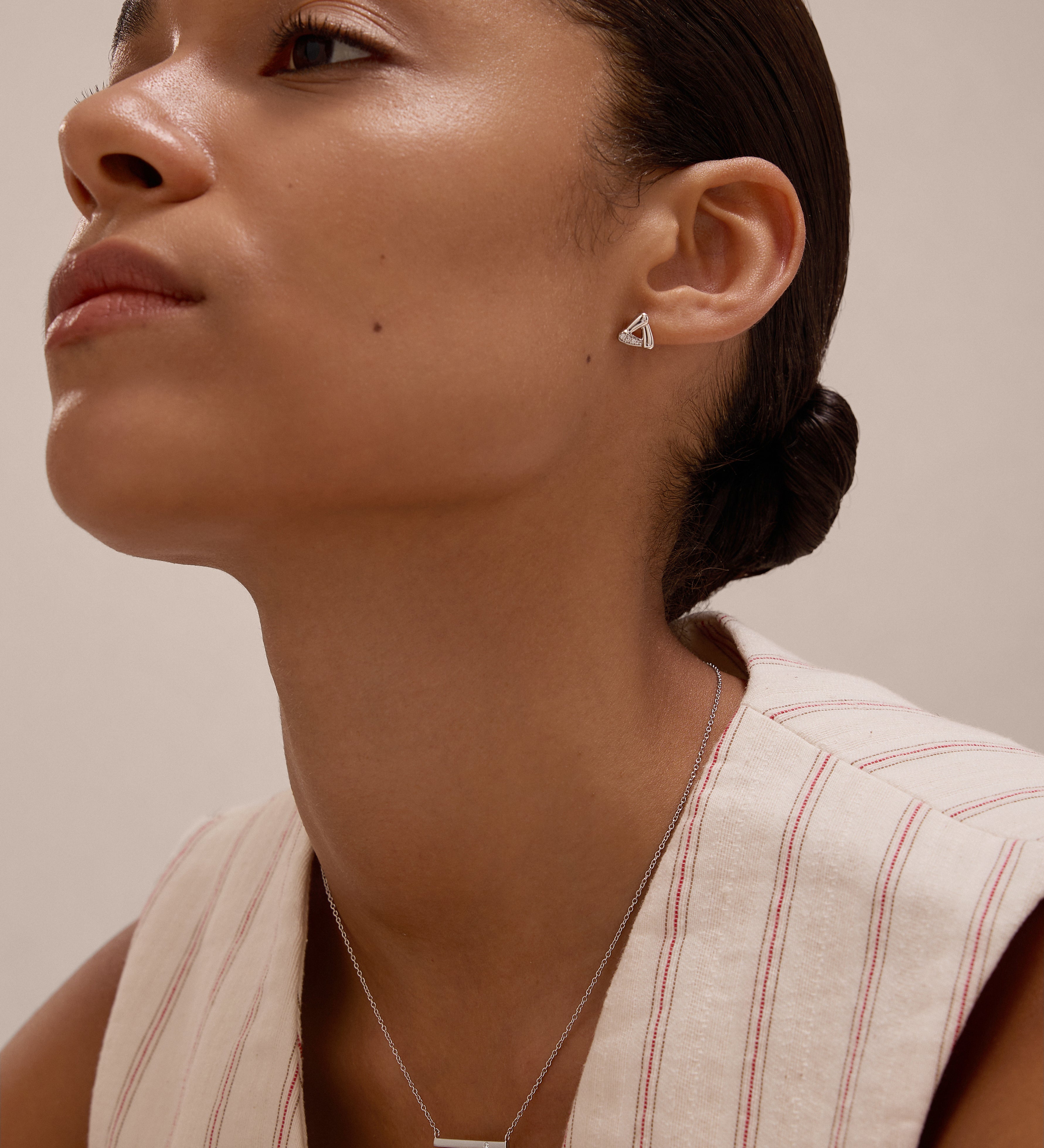 A woman with smooth skin and dark hair in a low bun wears a sleeveless cream top with red stripes, showcasing the Regina Horizontal Bar Pendant Necklace in Silver and minimalist triangle earrings as she gazes upward calmly.