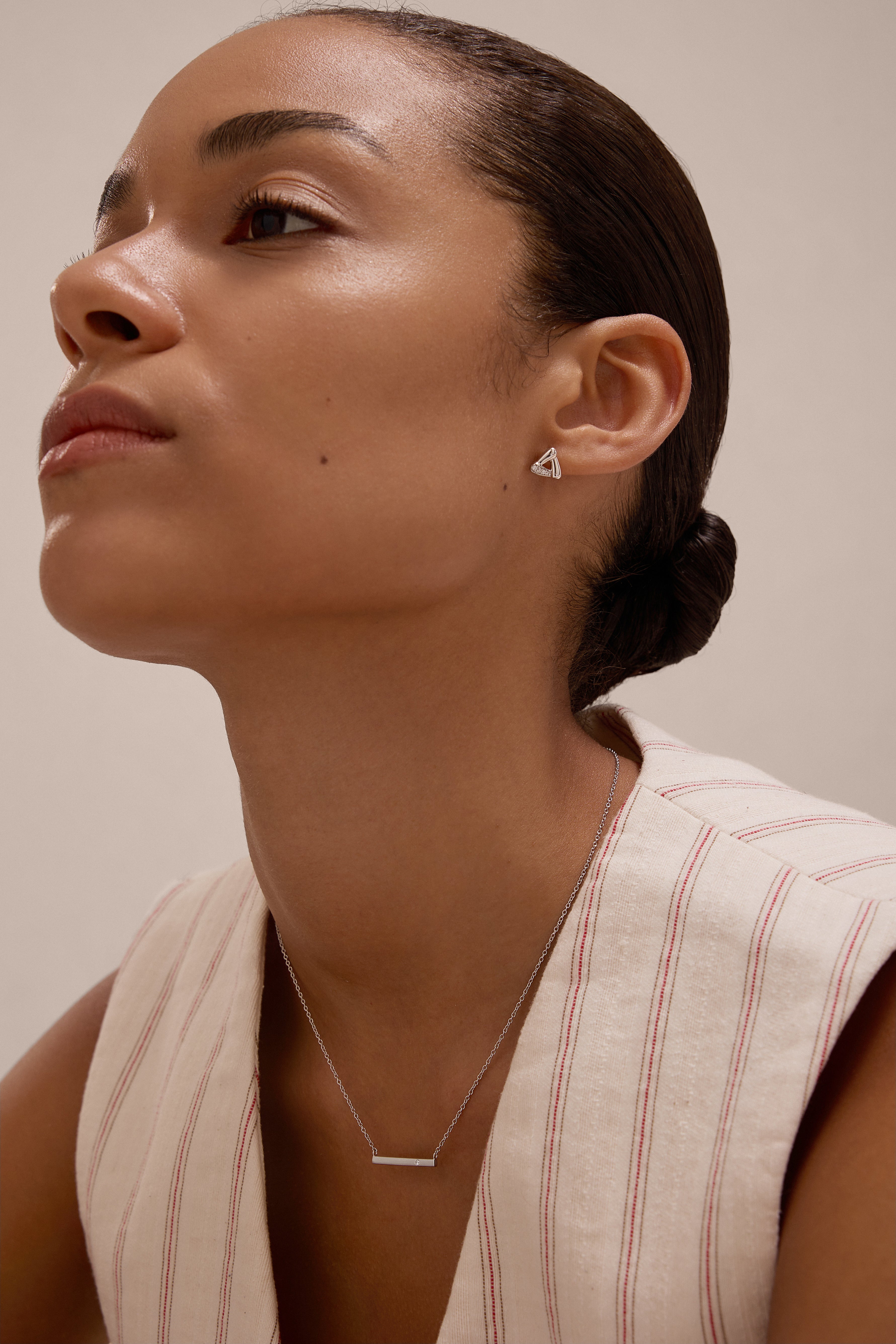 A woman with smooth skin and dark hair in a low bun wears a sleeveless cream top with red stripes, showcasing the Regina Horizontal Bar Pendant Necklace in Silver and minimalist triangle earrings as she gazes upward calmly.