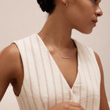 A woman with dark hair in a low bun wears a sleeveless cream top with red stripes and accessorizes with the Regina Horizontal Bar Pendant Necklace in Silver, embracing minimalist elegance against a light background.