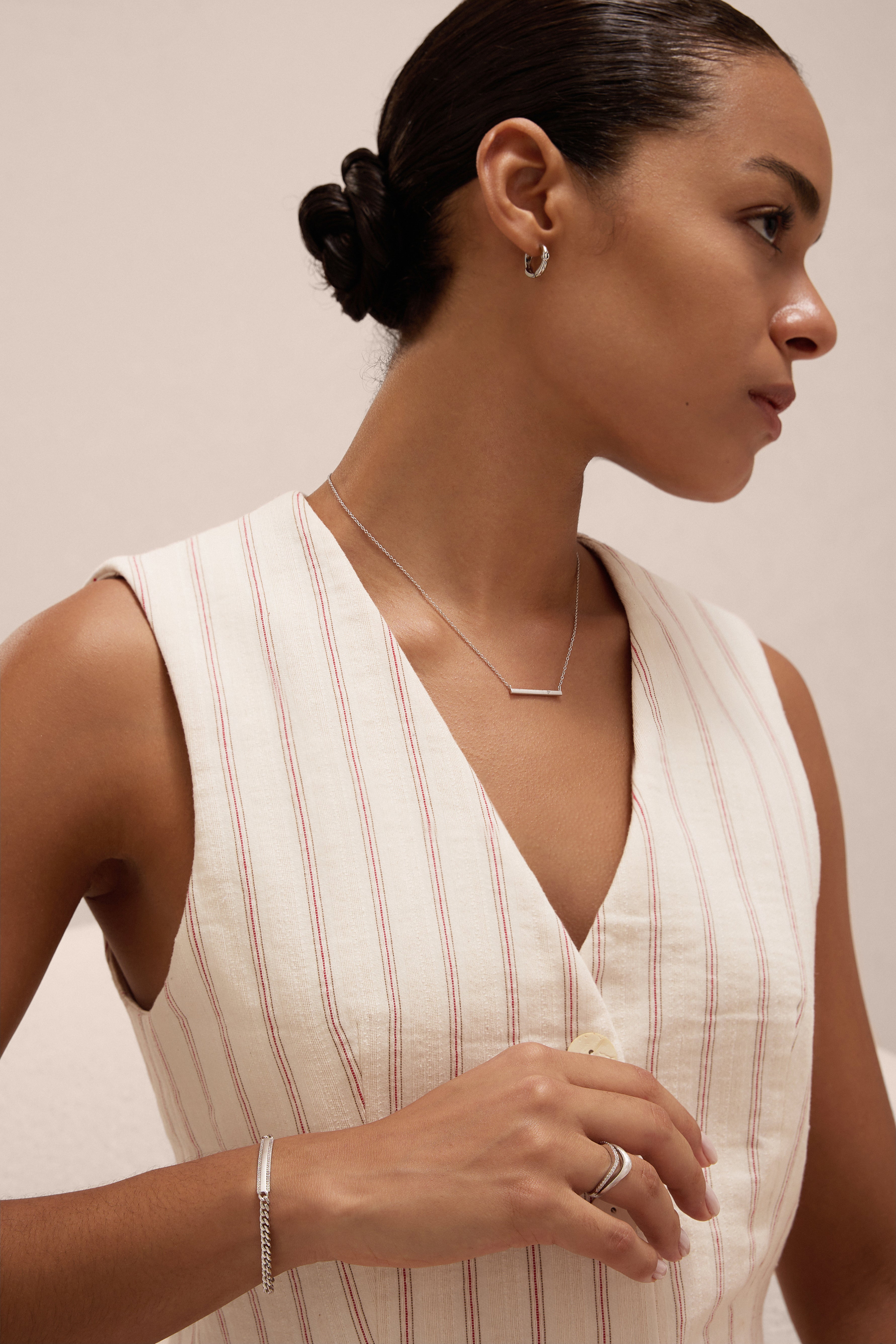A woman with dark hair in a low bun wears a sleeveless cream top with red stripes and accessorizes with the Regina Horizontal Bar Pendant Necklace in Silver, embracing minimalist elegance against a light background.