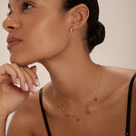 A woman with smooth, glowing skin and dark hair tied back wears the Thea Green Heart Pendant Gold Necklace with matching gold earrings. She poses in a black spaghetti strap top against a neutral background.