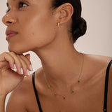 A woman with smooth, glowing skin and dark hair tied back wears the Thea Green Heart Pendant Gold Necklace with matching gold earrings. She poses in a black spaghetti strap top against a neutral background.