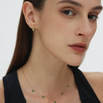A woman with straight brown hair wears a sleeveless black top, small green gemstone earrings, and the Thea Green Heart Pendant Gold Necklace. She looks slightly to the side with a neutral expression.