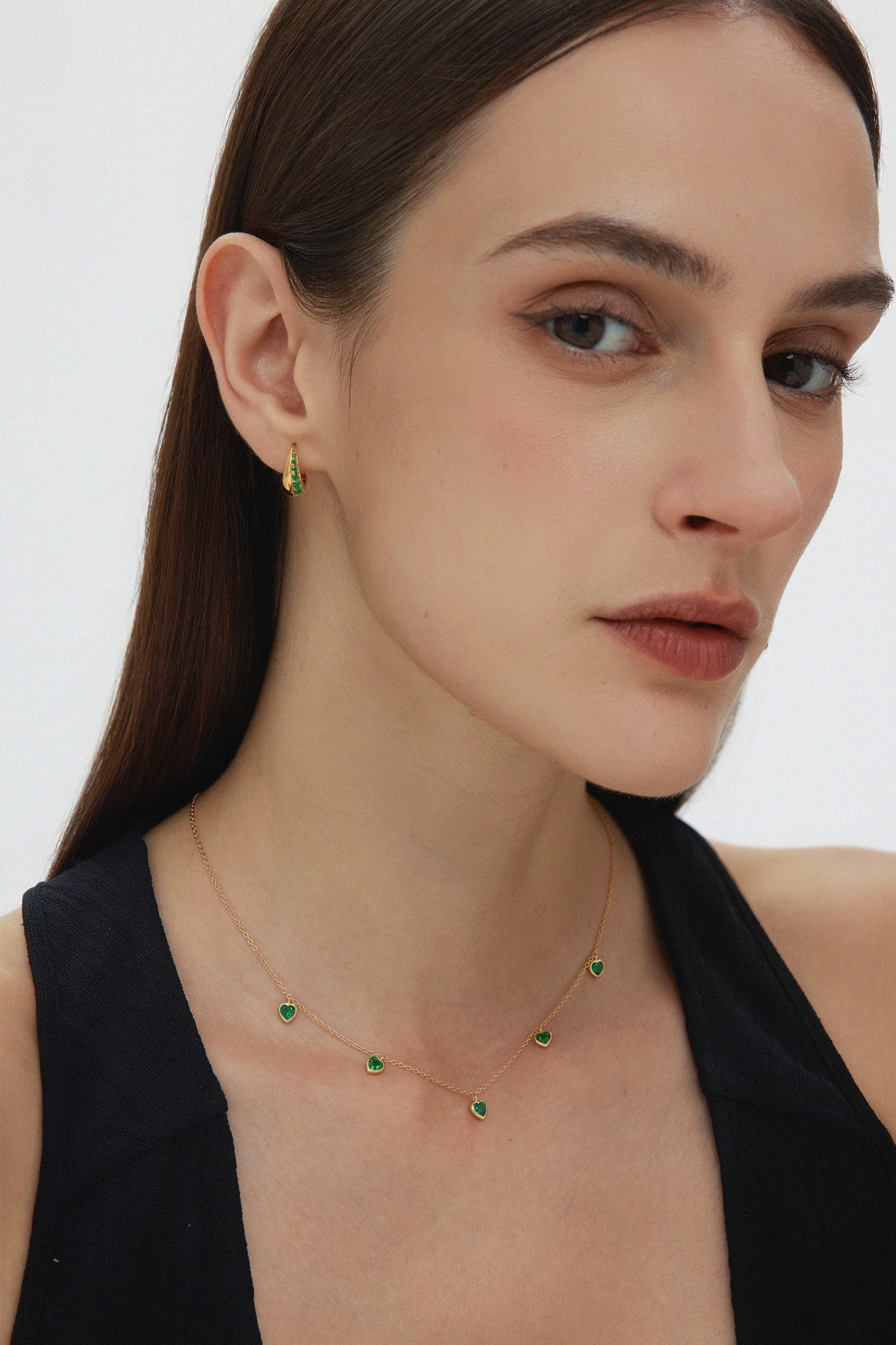 A woman with straight brown hair wears a sleeveless black top, small green gemstone earrings, and the Thea Green Heart Pendant Gold Necklace. She looks slightly to the side with a neutral expression.