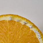 A close-up of a sliced orange displays the Marquise Threadless Flat Back Titanium Cartilage Stud in Gold, along with other sparkling gemstone earrings, arranged along the curved edge against a neutral background.
