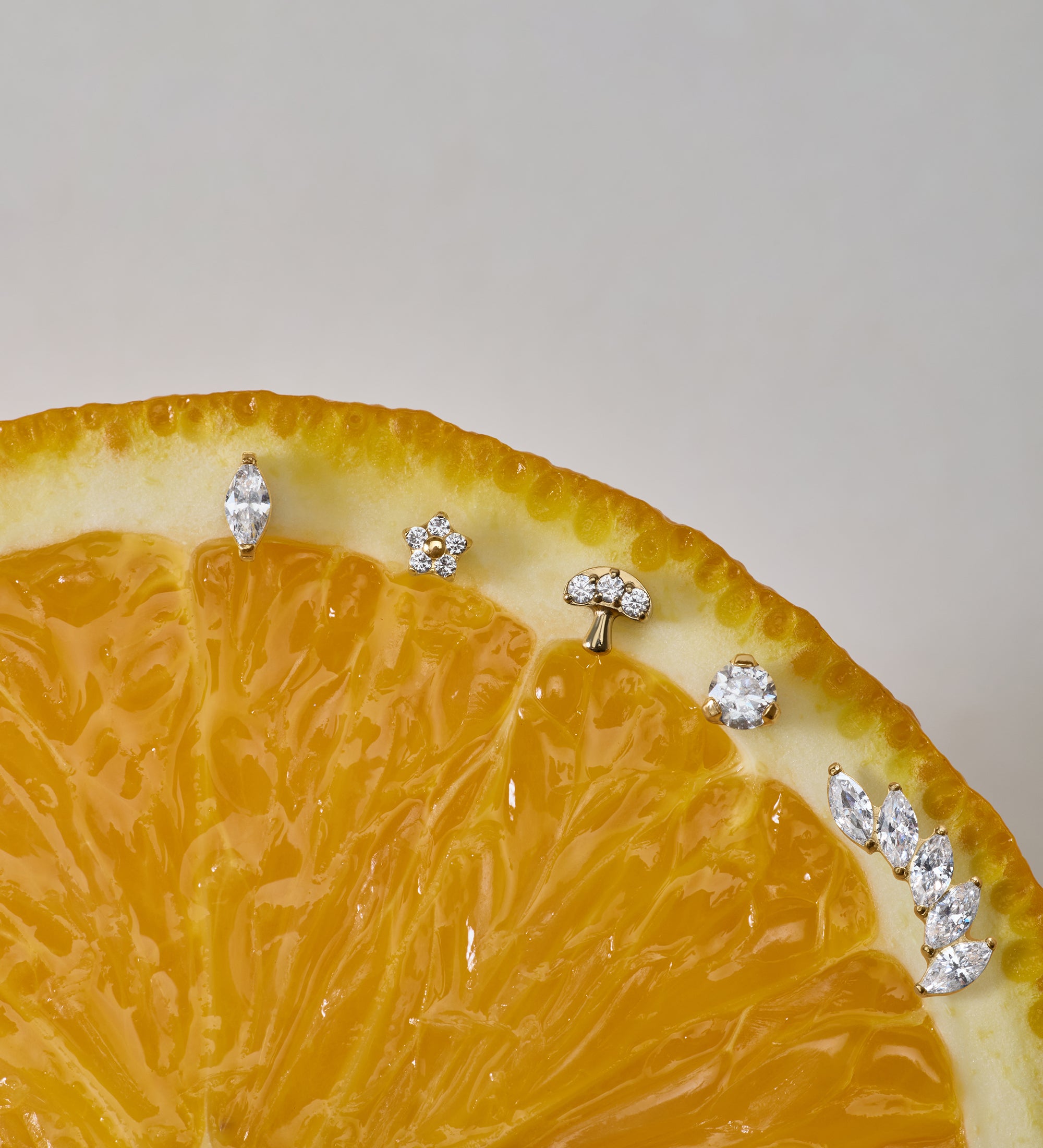 A close-up of a sliced orange displays the Marquise Threadless Flat Back Titanium Cartilage Stud in Gold, along with other sparkling gemstone earrings, arranged along the curved edge against a neutral background.