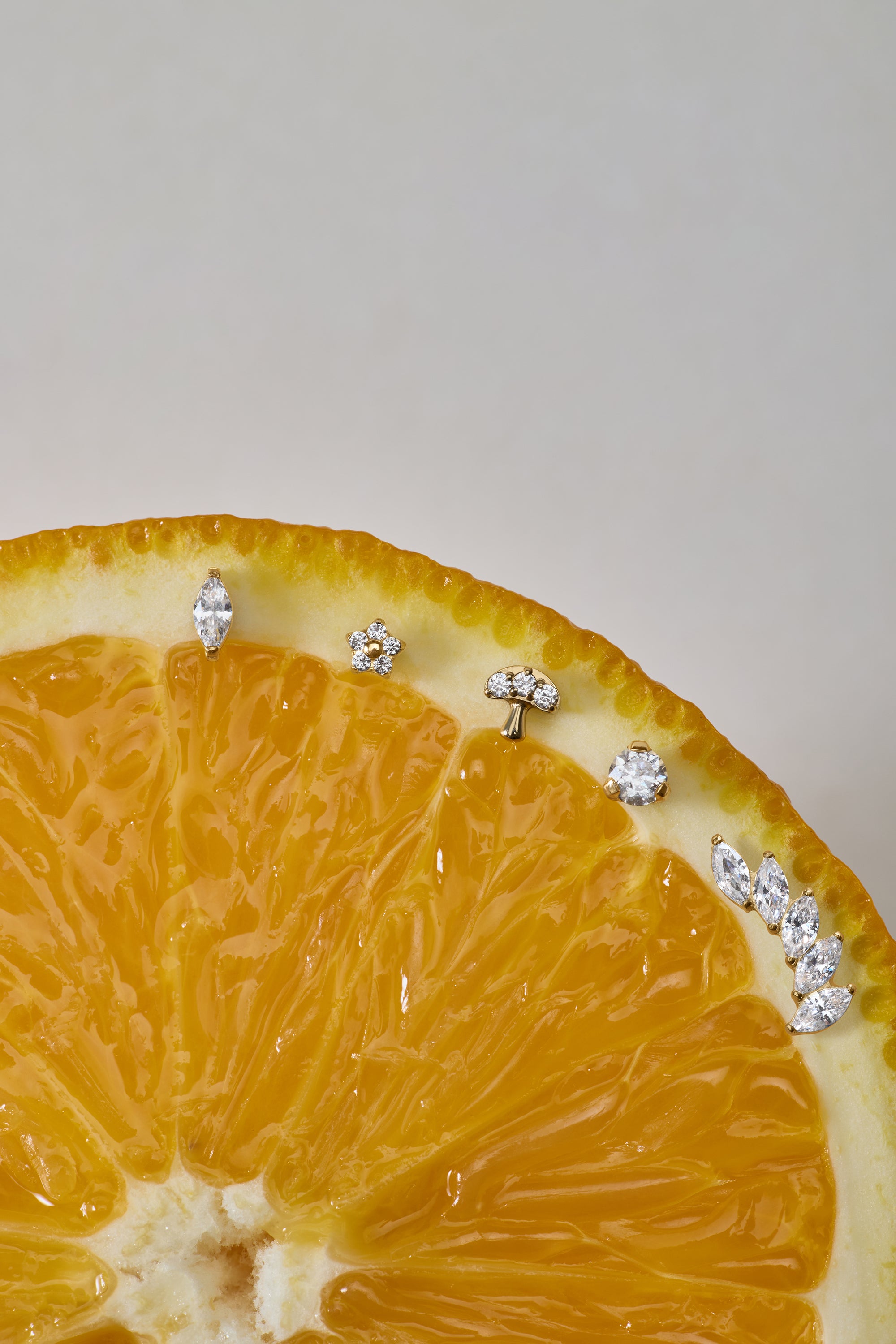A close-up of a sliced orange displays the Marquise Threadless Flat Back Titanium Cartilage Stud in Gold, along with other sparkling gemstone earrings, arranged along the curved edge against a neutral background.