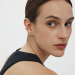 A woman with slicked-back dark hair and natural makeup wears a sleeveless black top, glancing over her shoulder. She sports the Vale Crystal Ear Cuff in Gold on her left ear, adding shimmer and sparkle against a plain white background.