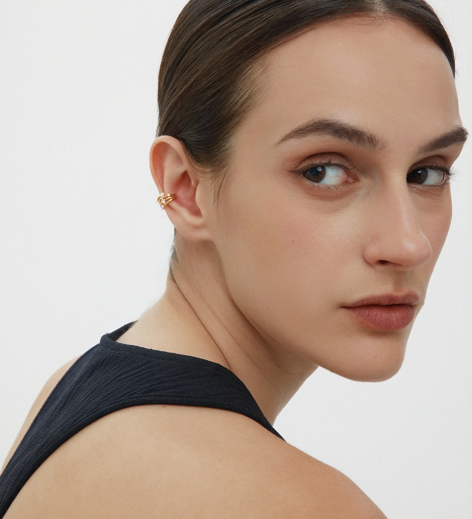 A woman with slicked-back dark hair and natural makeup wears a sleeveless black top, glancing over her shoulder. She sports the Vale Crystal Ear Cuff in Gold on her left ear, adding shimmer and sparkle against a plain white background.