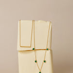 The Thea Green Heart Pendant Gold Necklace, featuring a small green cubic zirconia heart, is draped over two stacked blocks of butter against a neutral beige background.