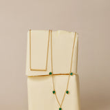 The Thea Green Heart Pendant Gold Necklace, featuring a small green cubic zirconia heart, is draped over two stacked blocks of butter against a neutral beige background.