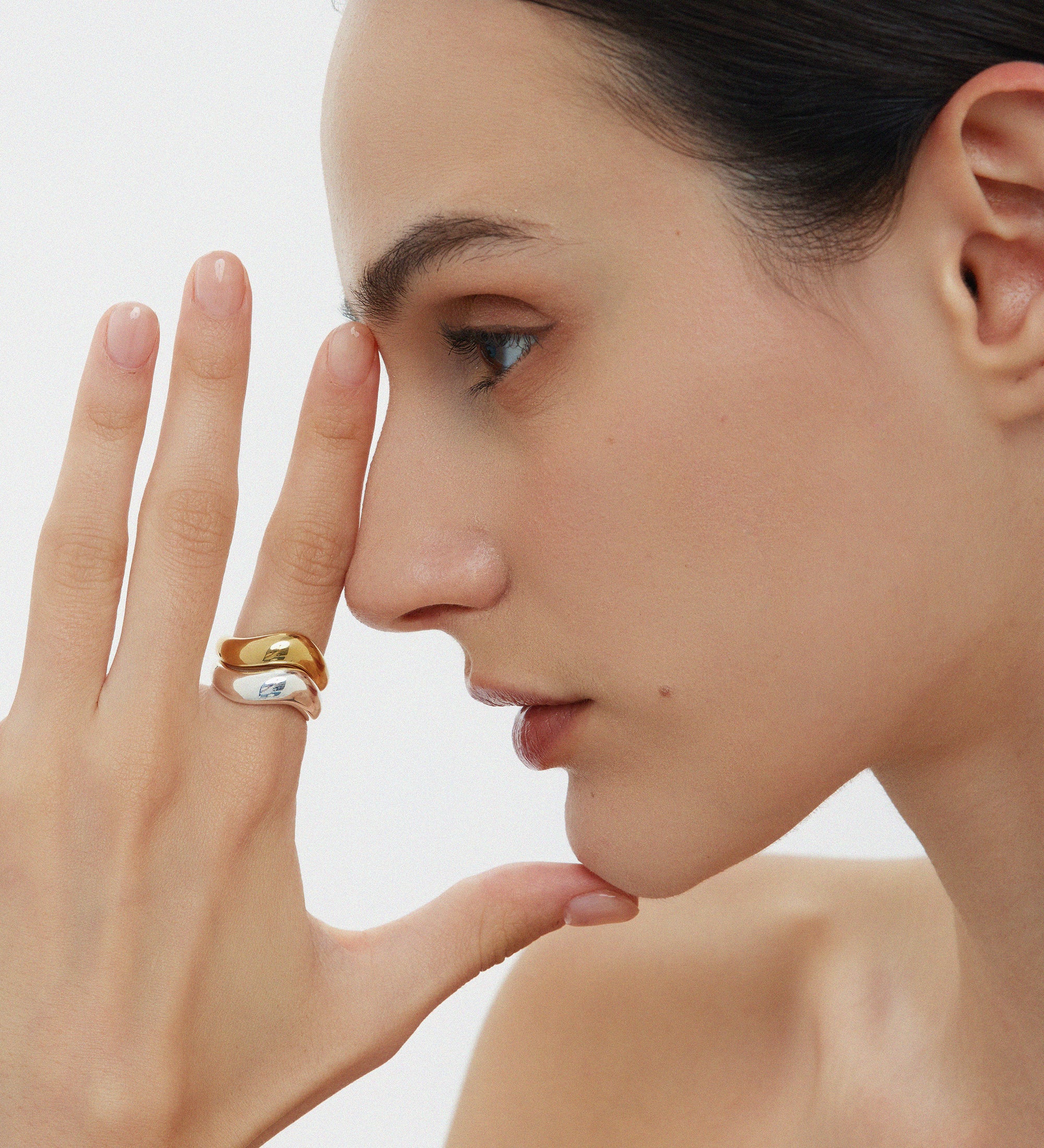 A woman with dark hair pulled back showcases the Vava Plain Chunky Curved Wave Ring in Gold, stacked with a minimalist gold-plated ring. She poses with a neutral expression against a plain background, her shoulders bare.
