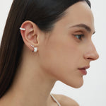 A woman with straight dark hair wears the Vera Twisted Crystal Ear Cuff in Silver on her upper ear, along with a small silver hoop in her lobe. She has natural makeup and is dressed in a white thin-strapped top.