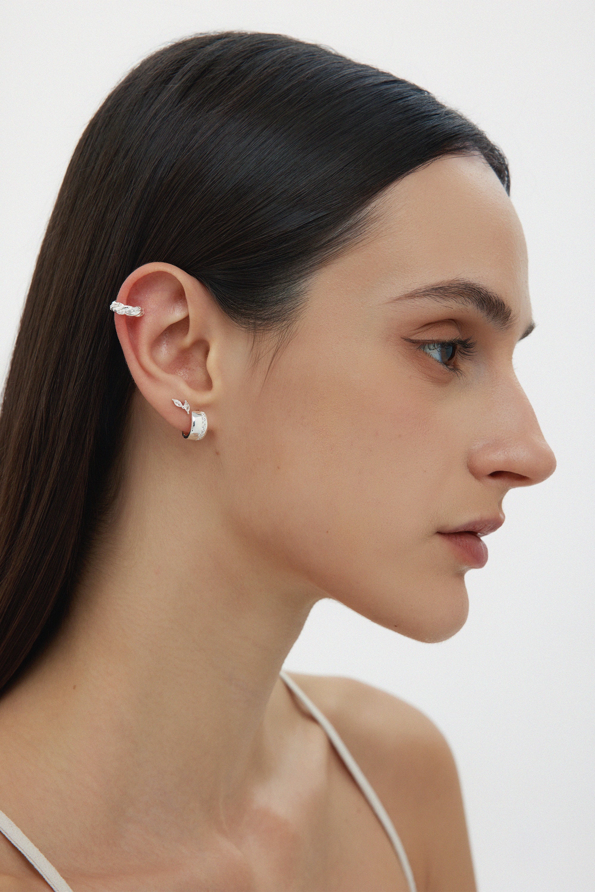 A woman with straight dark hair wears the Vera Twisted Crystal Ear Cuff in Silver on her upper ear, along with a small silver hoop in her lobe. She has natural makeup and is dressed in a white thin-strapped top.