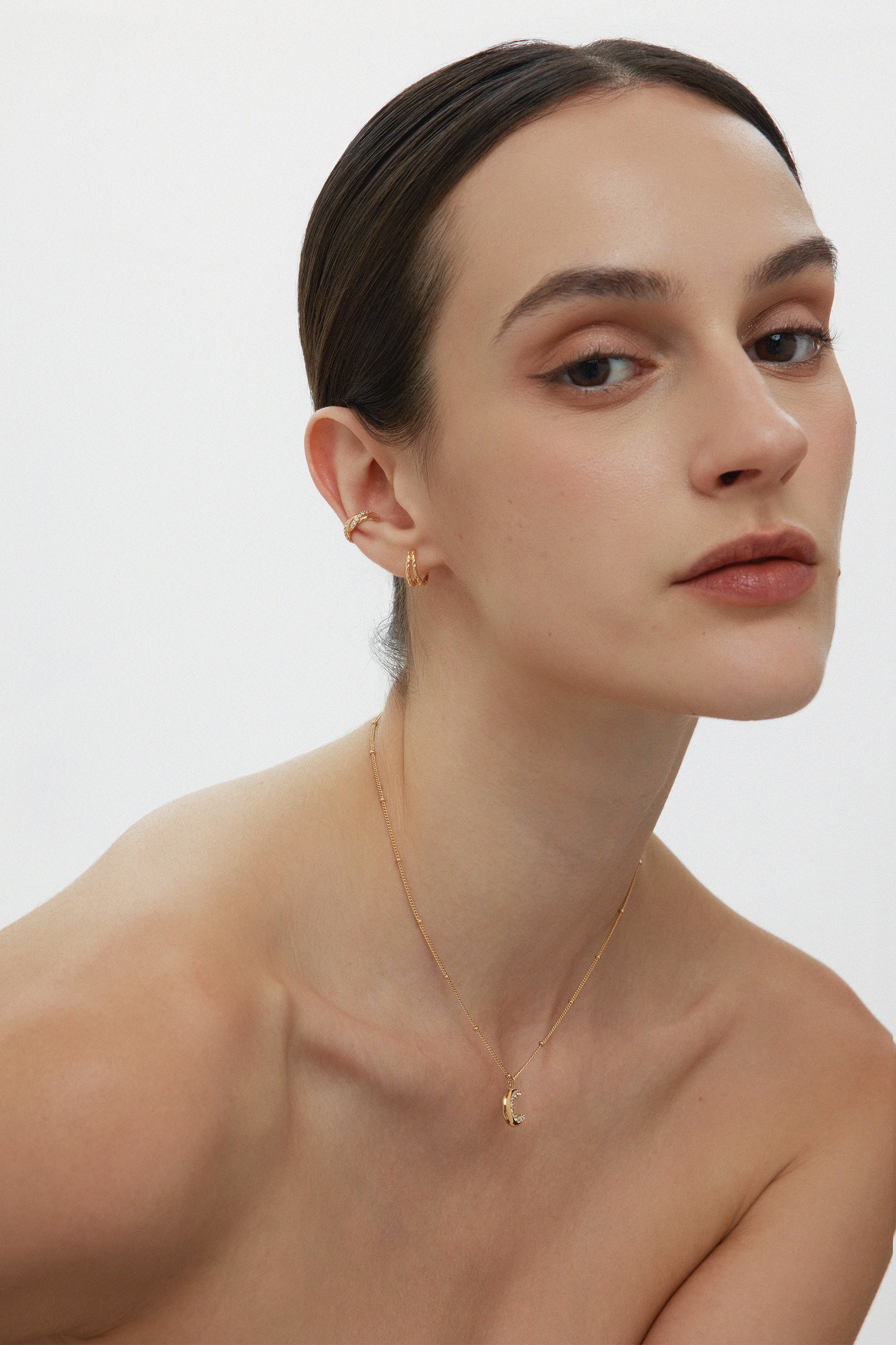 A woman with slicked-back hair wears small gold hoops, the Vera Twisted Crystal Ear Cuff in Gold, and a delicate pendant necklace, posing against a plain white background with a neutral expression.