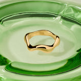 The Vica Fluid Wave Chunky Band Ring in Gold sits at the center of a smooth, modern clear green glass dish.