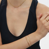 A woman in a sleeveless black top wears minimal gold jewelry—including the Vica Fluid Wave Chunky Band Ring in Gold—with her arms crossed and face partially out of frame against a plain background.