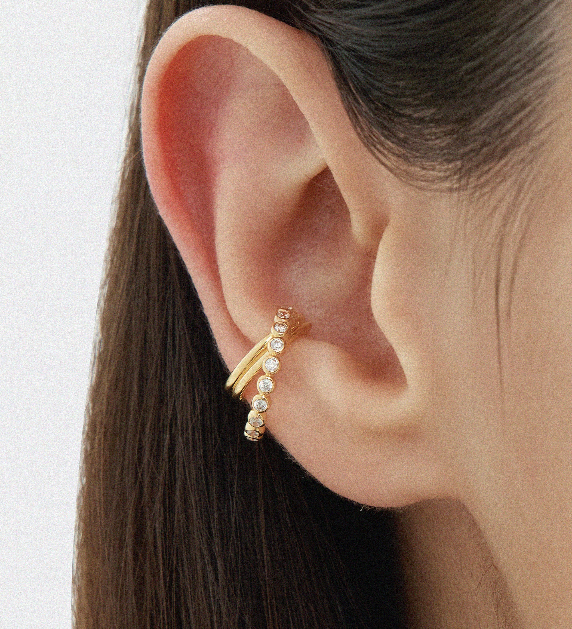Close-up of a persons ear wearing the Vina Crystal Bezel Ear Cuff in Gold, adorned with small round cubic zirconia. Straight brown hair frames the ear, set against a softly blurred background.