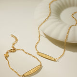 The Viva La Vida Bar Chain Bracelet in Gold, featuring an engraved bar and adjustable figaro chain, is displayed beside a white shell-shaped dish on a neutral background.
