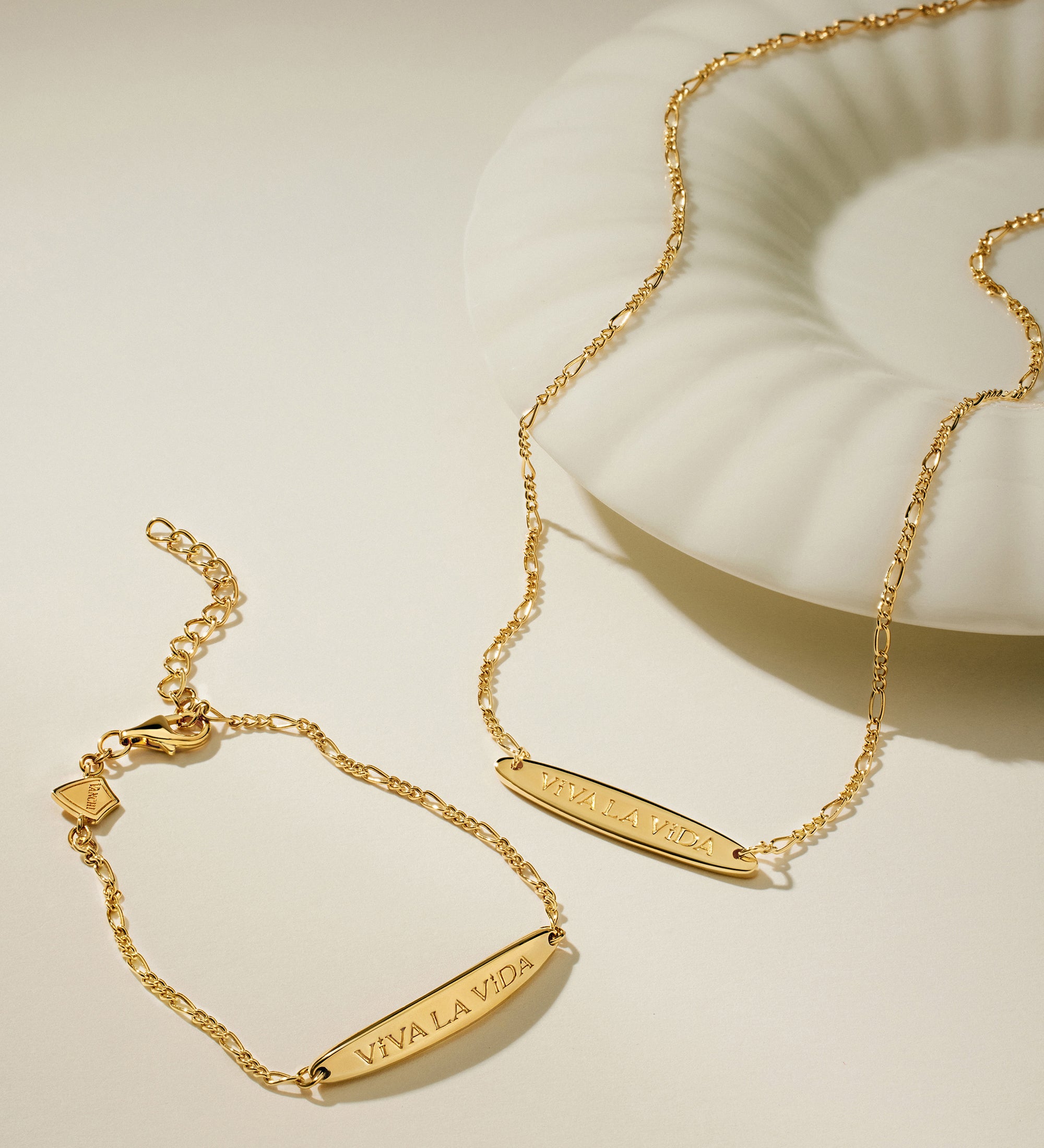 The Viva La Vida Bar Chain Necklace in Gold, featuring an adjustable figaro chain, rests alongside a matching gold bracelet on a light surface next to a white shell-shaped dish.