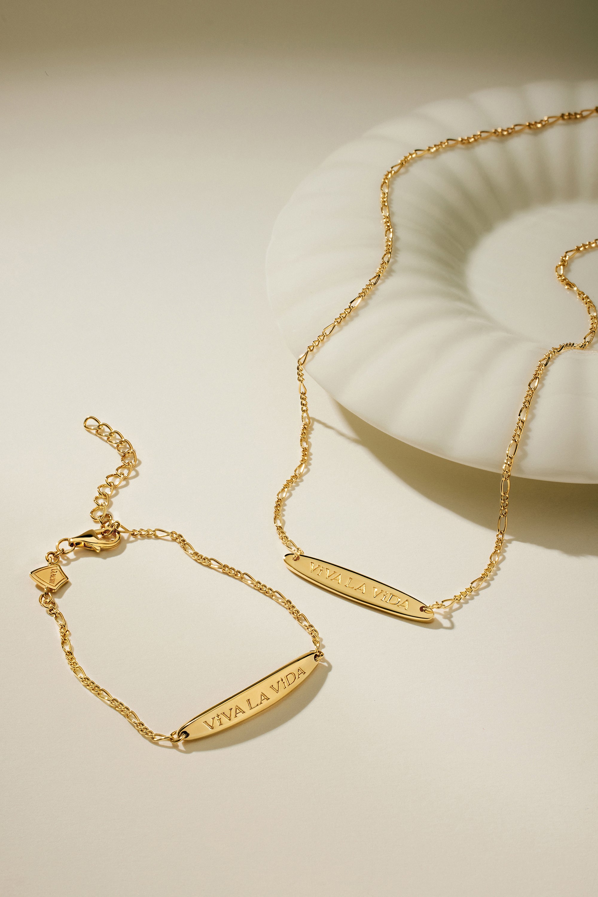 The Viva La Vida Bar Chain Necklace in Gold, featuring an adjustable figaro chain, rests alongside a matching gold bracelet on a light surface next to a white shell-shaped dish.