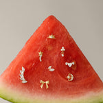 A slice of watermelon displays small gold and diamond earrings, including the Curved Marquise Threadless Flat Back Titanium Cartilage Stud in Platinum, set on its red flesh against a plain beige background.