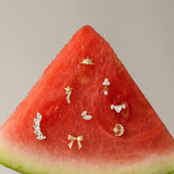 A slice of watermelon displays small gold and diamond earrings, including the Curved Marquise Threadless Flat Back Titanium Cartilage Stud in Platinum, set on its red flesh against a plain beige background.