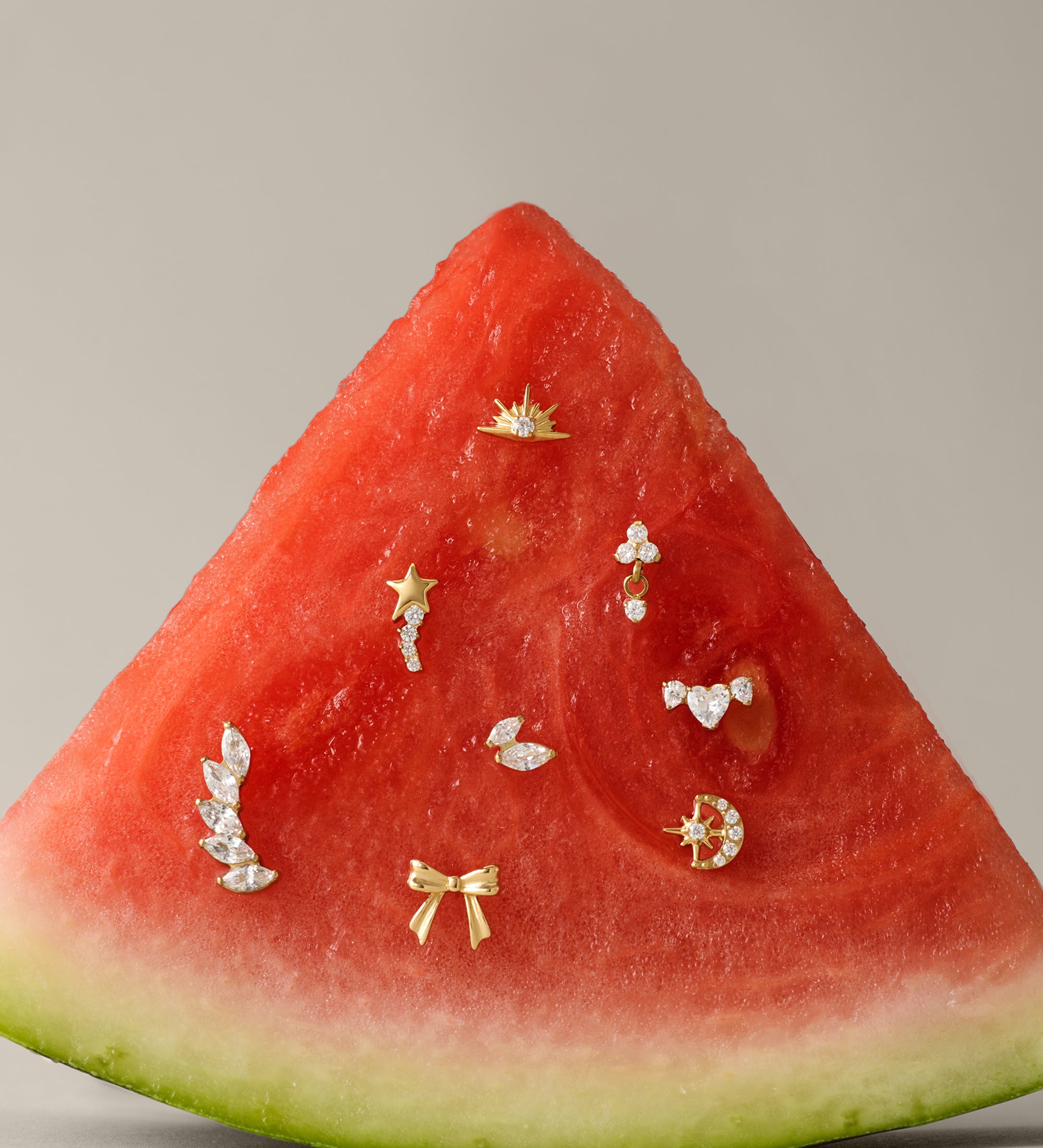 A slice of watermelon displays small gold and diamond earrings, including the Curved Marquise Threadless Flat Back Titanium Cartilage Stud in Platinum, set on its red flesh against a plain beige background.