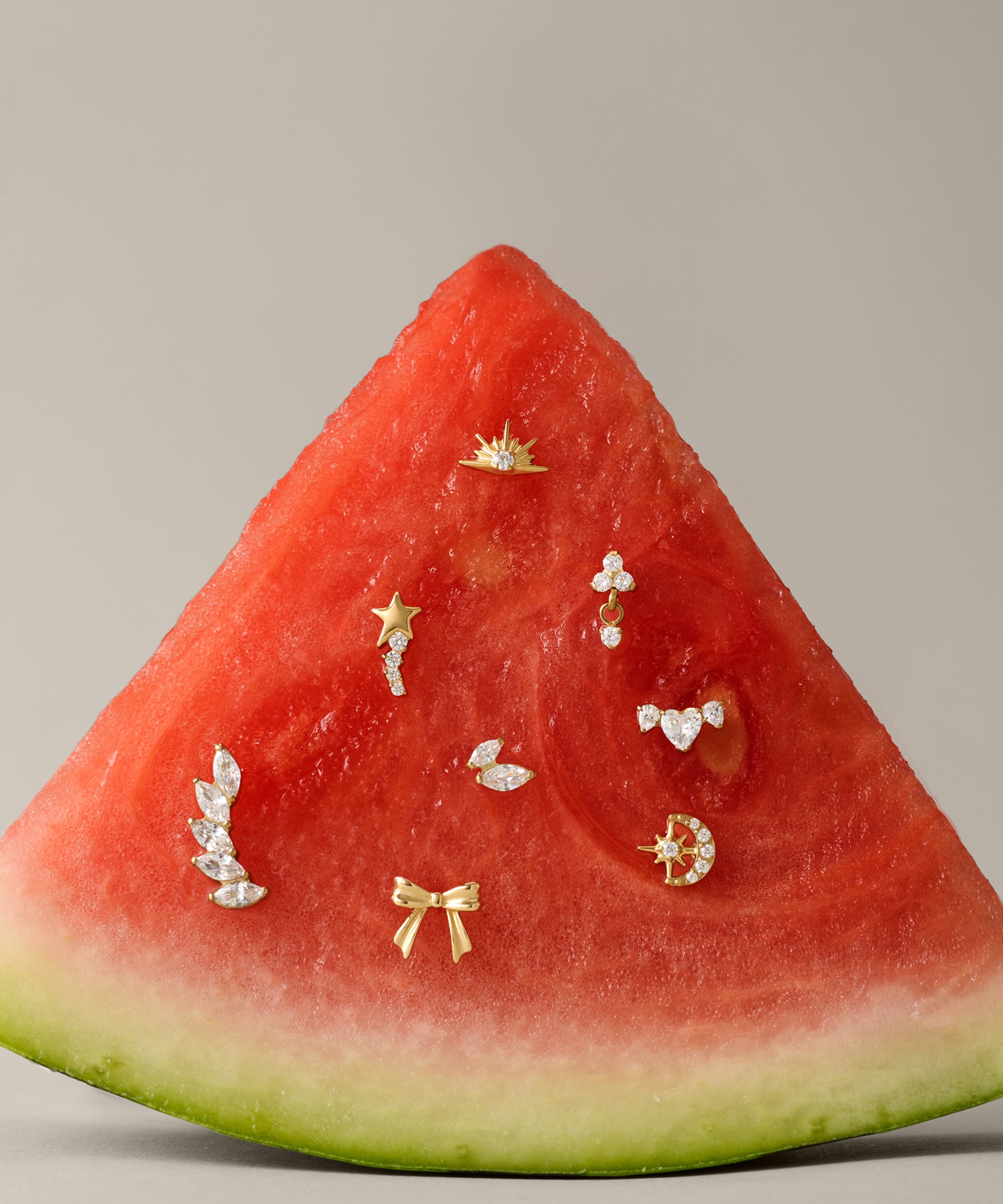 A slice of watermelon displays small gold and diamond earrings, including the Curved Marquise Threadless Flat Back Titanium Cartilage Stud in Platinum, set on its red flesh against a plain beige background.
