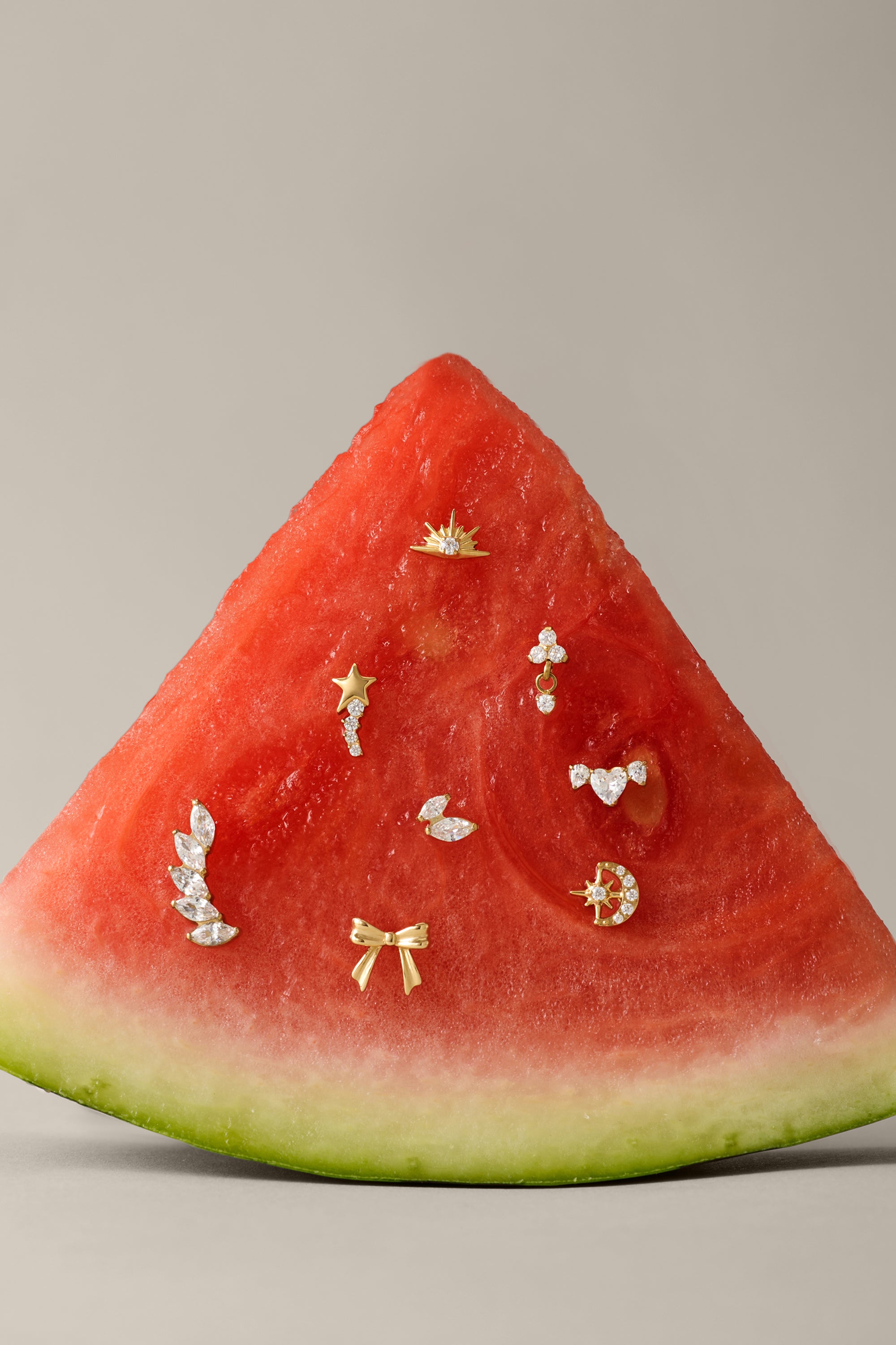 A slice of watermelon displays small gold and diamond earrings, including the Curved Marquise Threadless Flat Back Titanium Cartilage Stud in Platinum, set on its red flesh against a plain beige background.