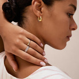 A woman with dark hair in a bun wears gold hoop earrings and the Yvette Two-Tone Mixed Metal Ring. She touches her neck with manicured fingers and is dressed in a white top featuring beige and red stripes.