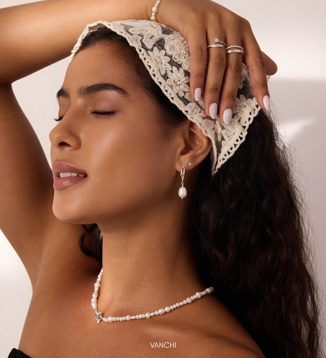 A woman with closed eyes wears a lace headscarf, pearl necklace, pearl earrings, rings, and a pearl bracelet. She poses with one hand touching her head, showcasing her jewelry along with the delicate 14K Solid White Gold Shell Flat Back Stud in her helix piercing. The background is neutral.