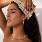 A woman with closed eyes wears a lace headscarf, pearl necklace, pearl earrings, rings, and a pearl bracelet. She poses with one hand touching her head, showcasing her jewelry along with the delicate 14K Solid White Gold Shell Flat Back Stud in her helix piercing. The background is neutral.